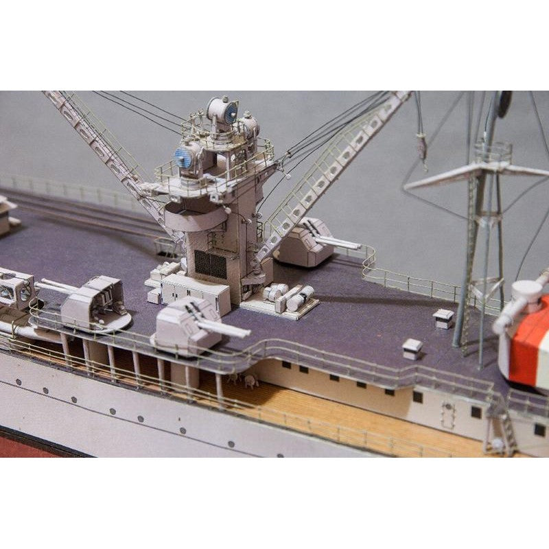 A close-up of a detailed model ship deck, showing gray and white gun turrets, antennas, cranes, and railings on a dark gray hull with a light wood deck edge.