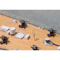 A close-up of a model ship's deck with several black and gray miniature weapon mounts, white crates, and a tan wood-textured surface against a gray background.