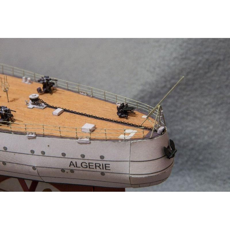 Close-up of a detailed model ship deck and hull, showing a beige wood-patterned deck, black chain, small black figures, and a white hull with the name ALGERIE printed on it.