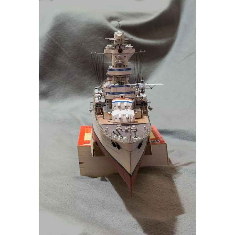 A detailed miniature model of a military battleship built from interlocking toy blocks, displayed on a small cardboard support against a draped gray fabric background.