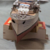 A close-up of a model ship or boat model photographed indoors, resting on small orange-and-white blocks against a gray surface.