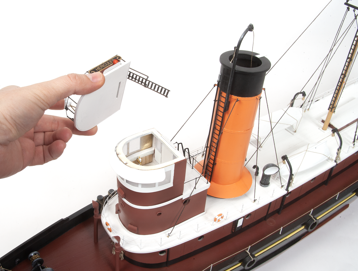 A hand holding a small white accessory piece next to a detailed model ship with a brown hull, white deck, and orange smokestack on a white background.