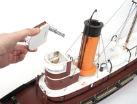 A hand holding a small white accessory piece next to a detailed model ship with a brown hull, white deck, and orange smokestack on a white background.