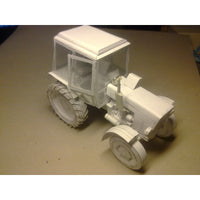 A white unpainted card model tractor with a cabin, photographed on a plain tan surface.