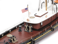 A close-up of a detailed model tugboat, featuring a dark red and white hull, a white deck, a small American flag, and realistic nautical accessories like ropes and cleats, set against a plain white background.