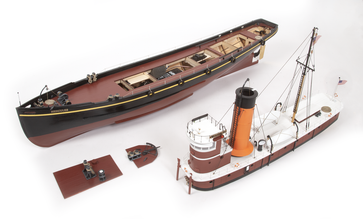 A detailed model ship set on a white background, featuring a long dark brown and black vessel, a smaller white and orange tugboat, and several small accessories.