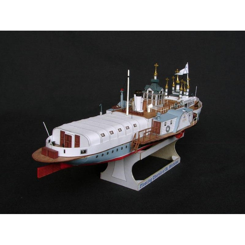 A detailed model of a passenger ship, likely a toy or scale model, displayed on a stand against a solid black background. The ship features a white upper deck, a light blue hull, brown wooden-style decks and railings, and small gold accents on the antennas and masts.