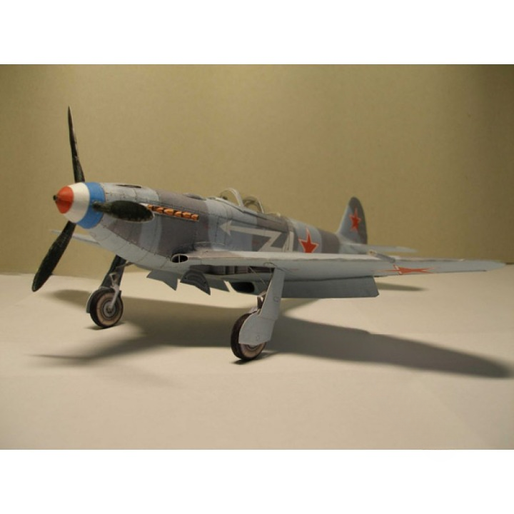 A model of a Soviet fighter plane, likely a MiG-15, painted in a gray camouflage scheme with red star insignias, orange text, and a blue and red nose cone, displayed on a neutral beige background.