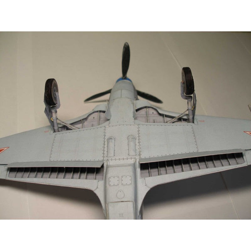 A detailed model airplane viewed from the underside, featuring a light gray body, black propeller blades, and small red insignia on the wingtips, set against a plain beige background.