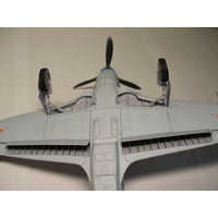 A detailed model airplane viewed from the underside, featuring a light gray body, black propeller blades, and small red insignia on the wingtips, set against a plain beige background.