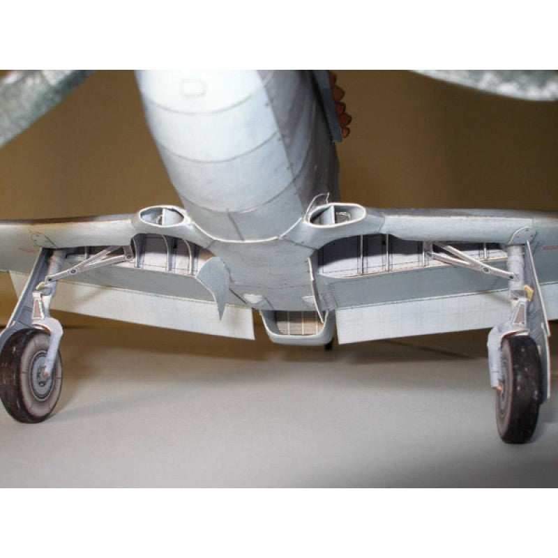 Close-up underside of a model airplane showing the landing gear, wing struts, and fuselage details against a plain indoor background.