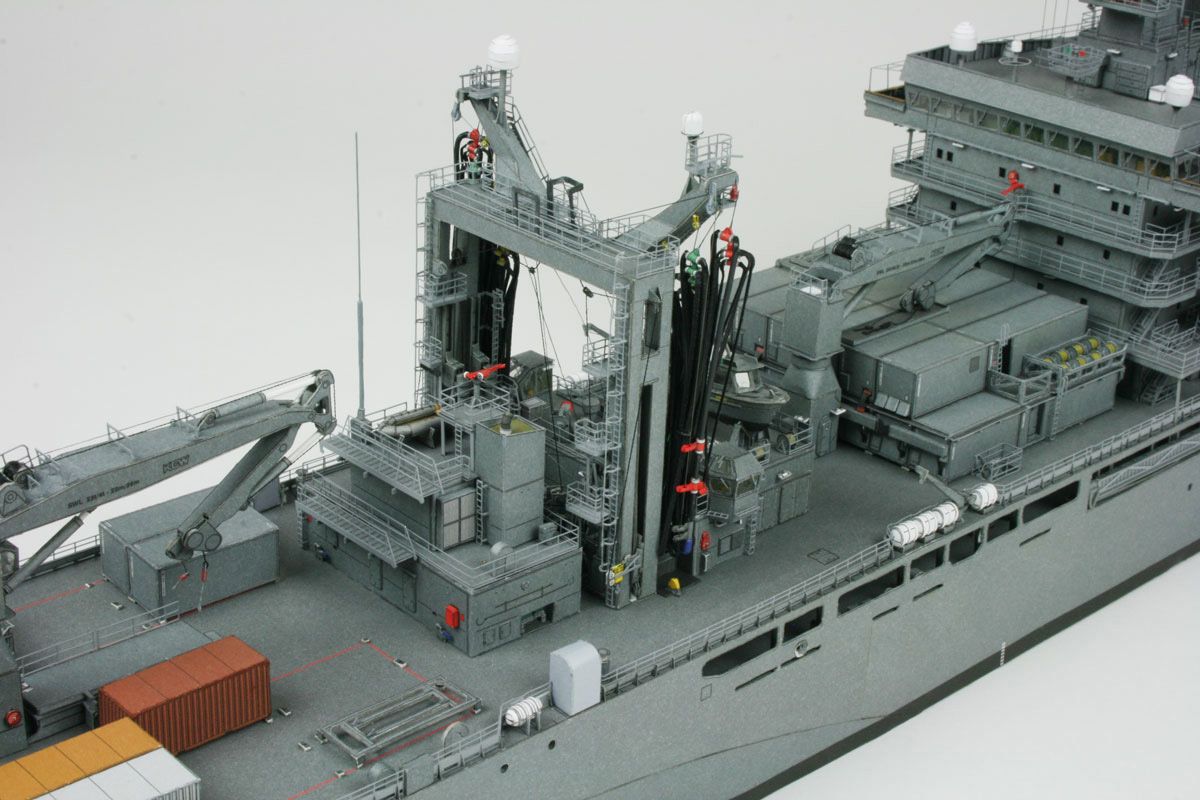 A detailed model of a gray military cargo ship deck, featuring complex machinery, cranes, antennas, and small red and orange accents, photographed against a plain light gray background.
