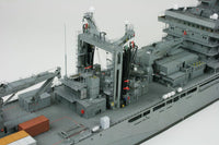 A detailed model of a gray military cargo ship deck, featuring complex machinery, cranes, antennas, and small red and orange accents, photographed against a plain light gray background.