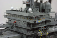 A close-up of a detailed gray model ship or aircraft carrier deck structure with multiple decks, railings, antennas, and small yellow and red accents, photographed against a plain light background.