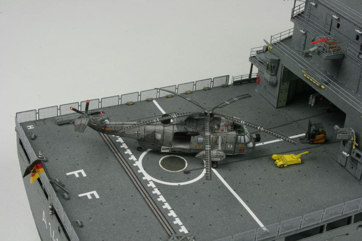 A detailed model of a military helicopter on a gray aircraft carrier deck, shown from an elevated perspective against a light gray background.
