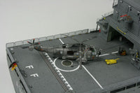 A detailed model of a military helicopter on a gray aircraft carrier deck, shown from an elevated perspective against a light gray background.