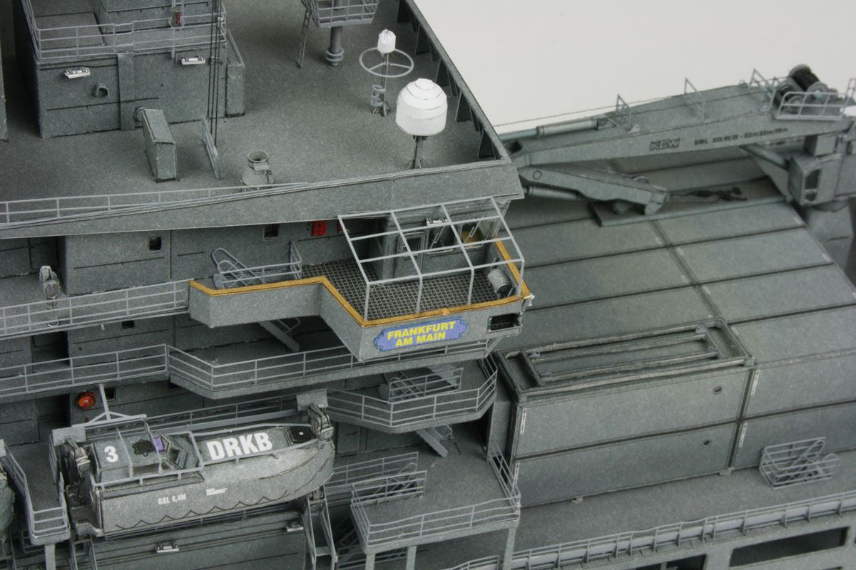 Close-up detail of a gray military ship model or miniature, showing decks, stairways, antennas, and a small lifeboat with DRKB markings.