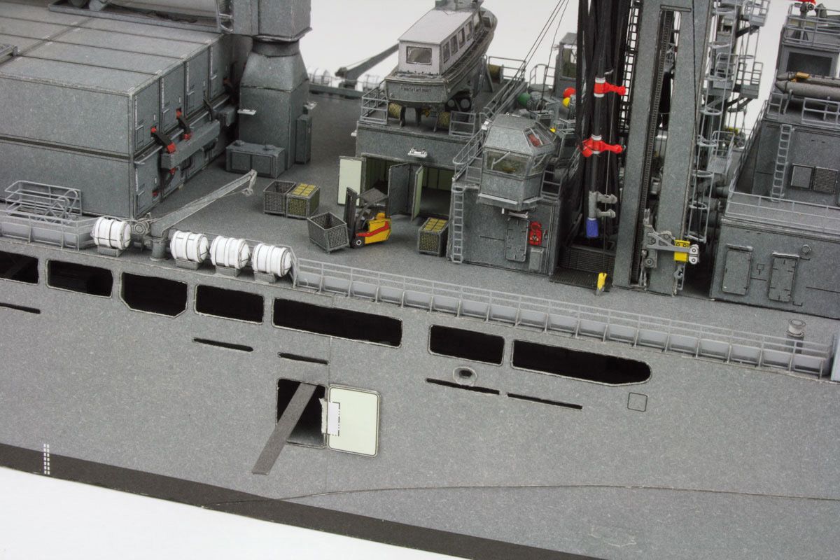 Close-up detail of a gray military ship deck with antennas, equipment, railings, and a small yellow forklift model.