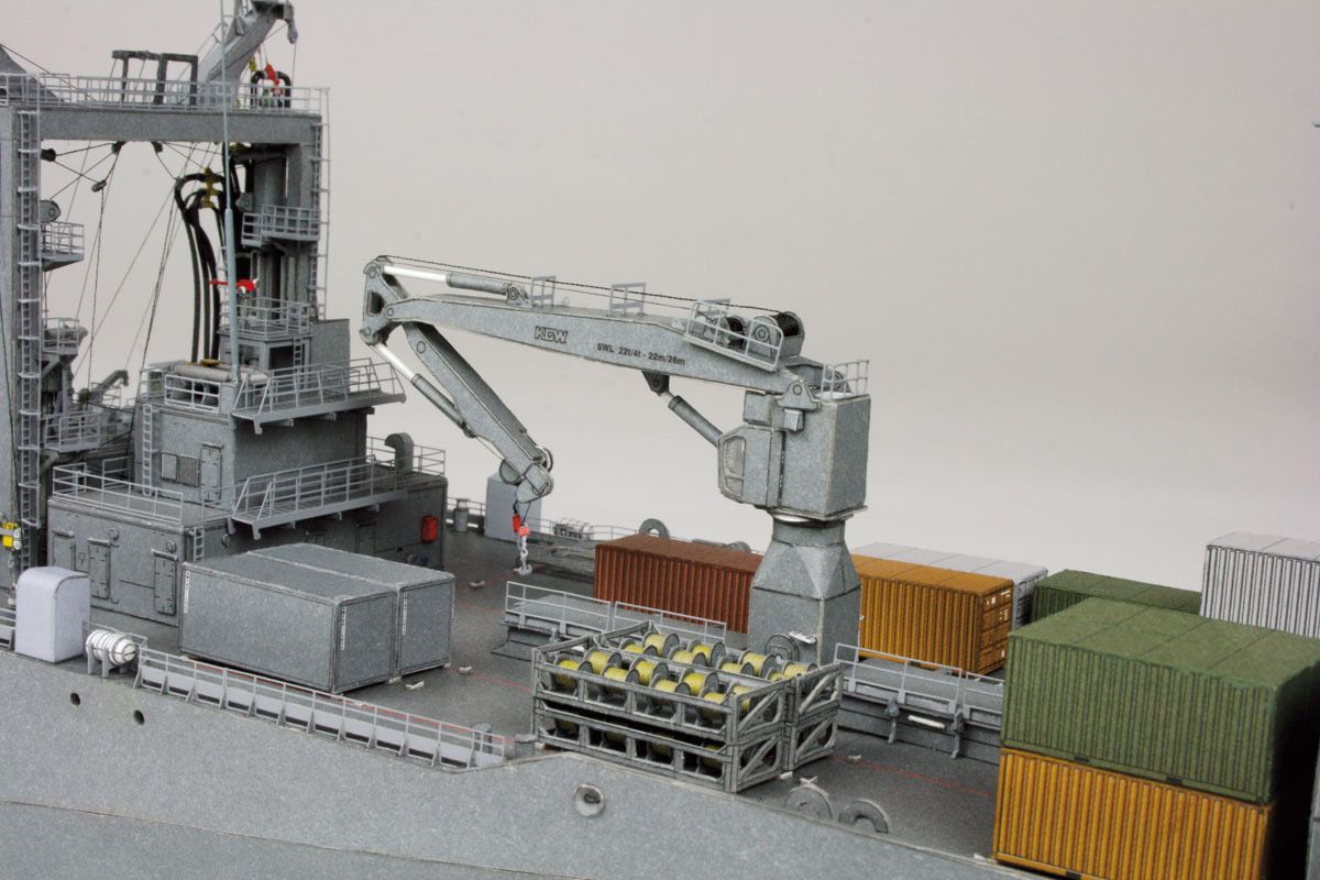 A detailed model of a cargo ship deck featuring a gray gantry crane, stacked shipping containers in green, orange, and white, and a gray superstructure, set against a plain light gray background.