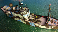 A small model fishing vessel named 'Steve Sawyer 77' painted in a blue, gray, and brown camouflage pattern, floating on green-blue water.