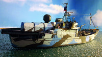 A small-scale model of a military survey boat with a blue, green, and brown camouflage pattern, floating on water under a blue sky with clouds.