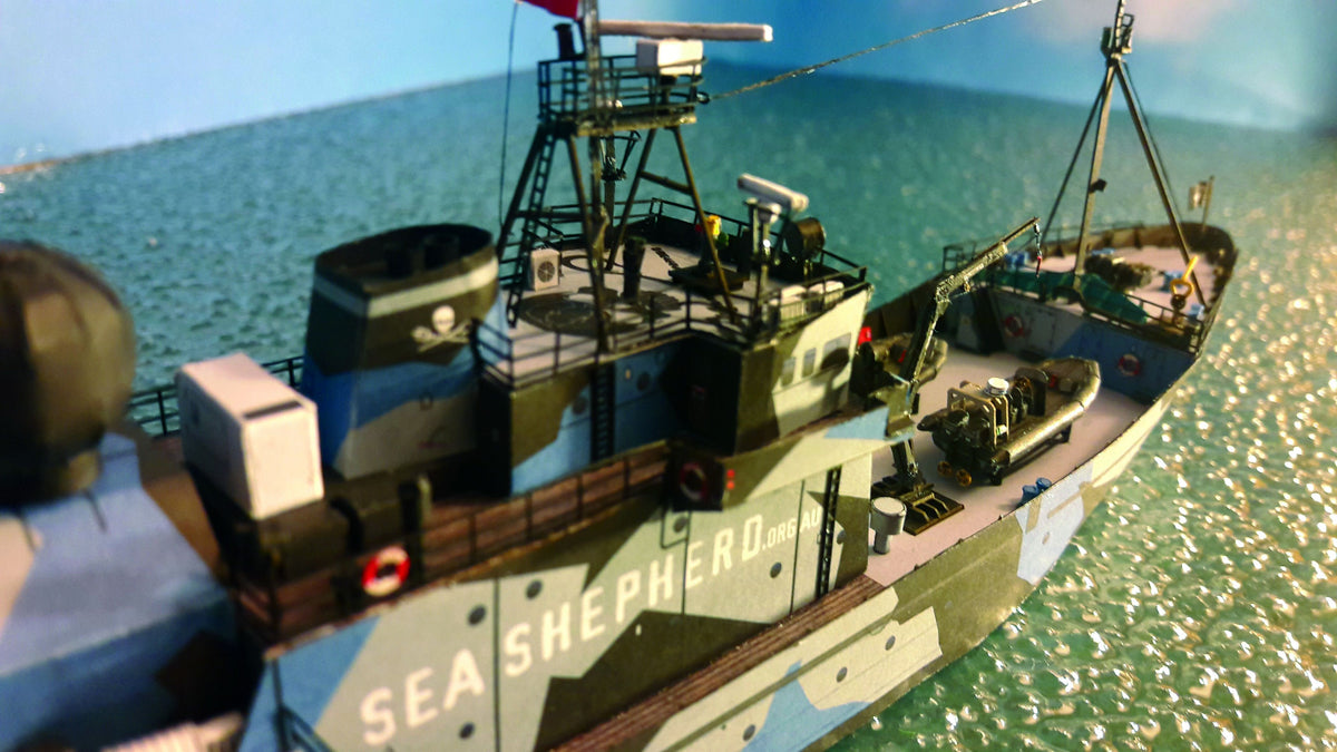 A close-up shot of a model fishing vessel named 'Sea Shepherd 005' with a green, gray, and black camouflage pattern, set against a simulated ocean background.