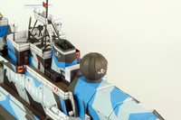 Close-up of a model ship or boat section with blue, white, and gray camouflage, black deck details, and a dark gray spherical radar dome on a plain light background.