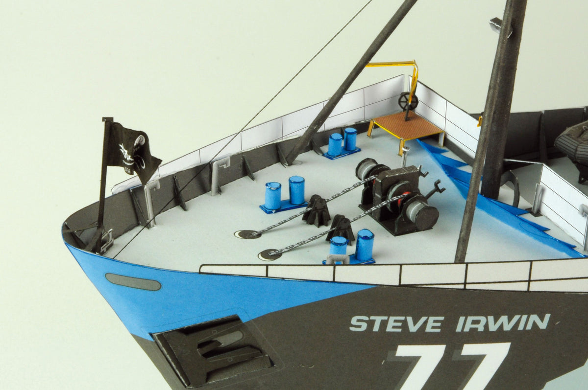 Close-up of a model ship deck labeled 'STEVE IRWIN' and '77', showing blue and black hull, white deck, winches, ropes, and a small yellow table against a plain light background.