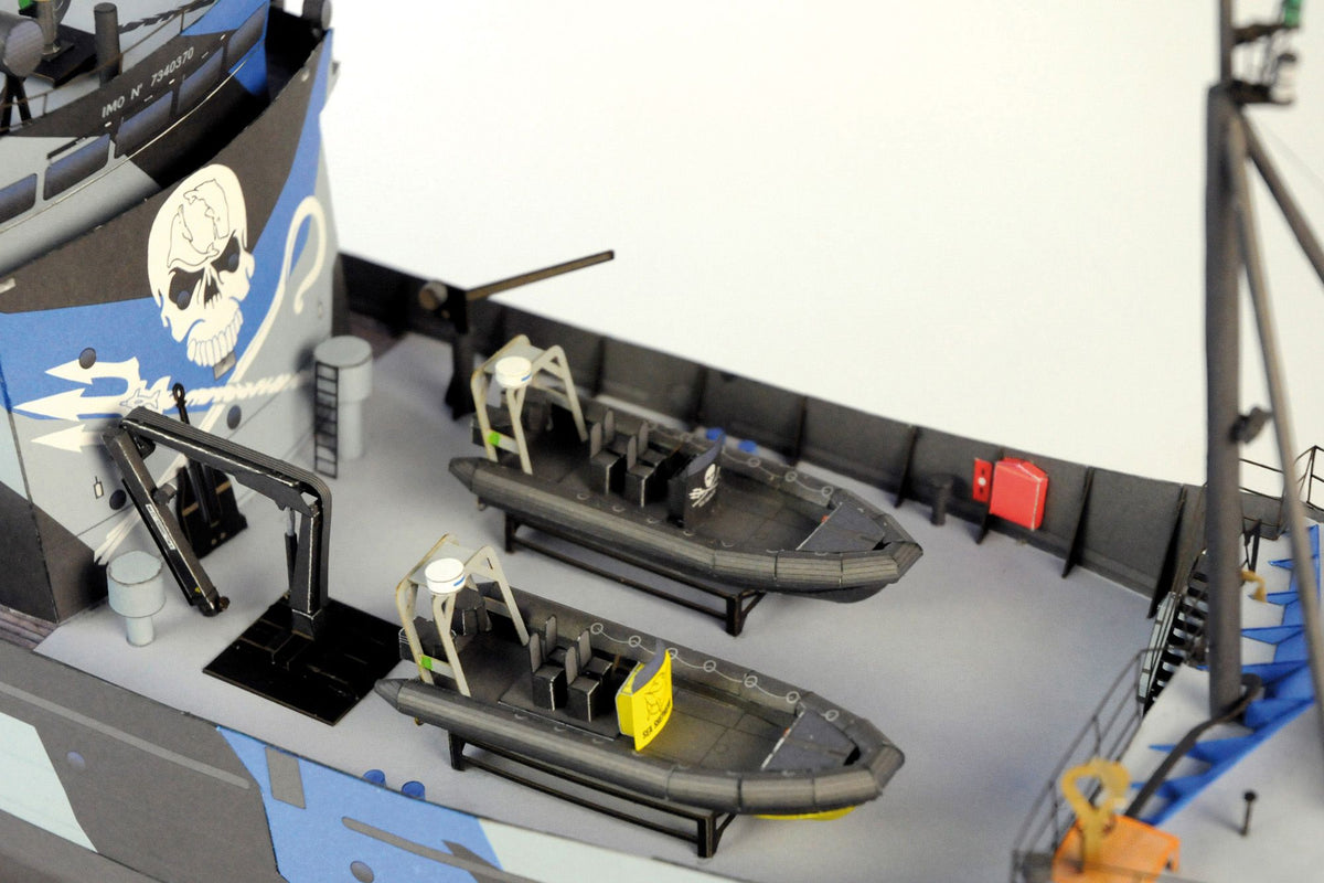 A close-up of a model ship deck featuring two gray inflatable boats with yellow life rafts, a blue and white painted hull, and black deck equipment on a white background.