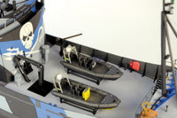 A close-up of a model ship deck featuring two gray inflatable boats with yellow life rafts, a blue and white painted hull, and black deck equipment on a white background.
