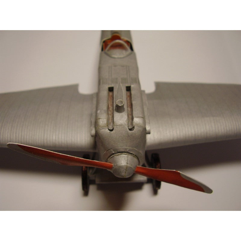 Close-up of a model airplane nose section with a silver-gray fuselage, wings, and red propeller blades against a plain light background.