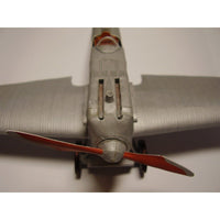 Close-up of a model airplane nose section with a silver-gray fuselage, wings, and red propeller blades against a plain light background.