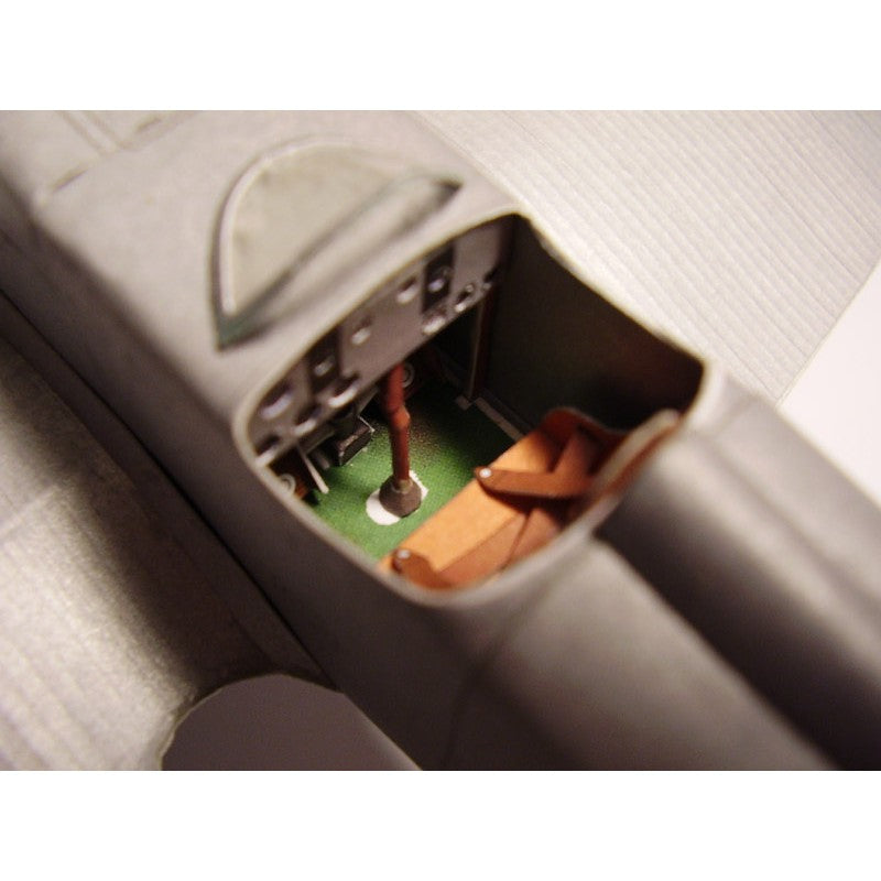 Close-up of a miniature model airplane cockpit interior, showing a green floor, brown seats, and gray fuselage with a clear windshield.