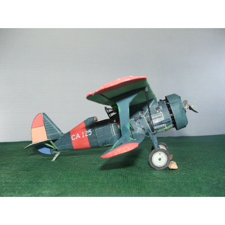 A vintage biplane model airplane with a dark teal body, red wingtips, and a red-and-orange tail, displayed on a green textured surface against a light gray background.
