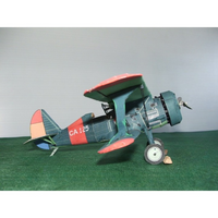 A vintage biplane model airplane with a dark teal body, red wingtips, and a red-and-orange tail, displayed on a green textured surface against a light gray background.