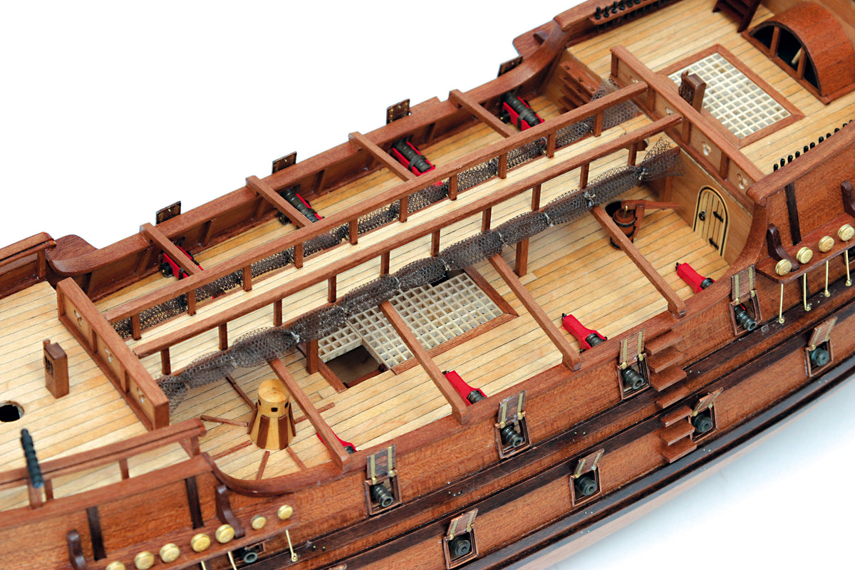 Close-up of a detailed wooden model ship, showing the deck, railings, cabin details, and small red accents on a white background.