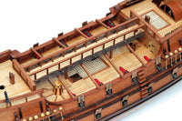Close-up of a detailed wooden model ship, showing the deck, railings, cabin details, and small red accents on a white background.