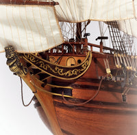 Close-up of a wooden model or replica ship's bow with white sails, dark brown hull, gold decorative trim, and a black-and-gold figure at the quarter.