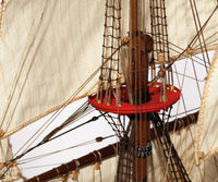 Close-up detail of a wooden model ship's mast and rigging, featuring a red horizontal crossbar, dark brown wooden poles, and cream-colored canvas sails against a white background.