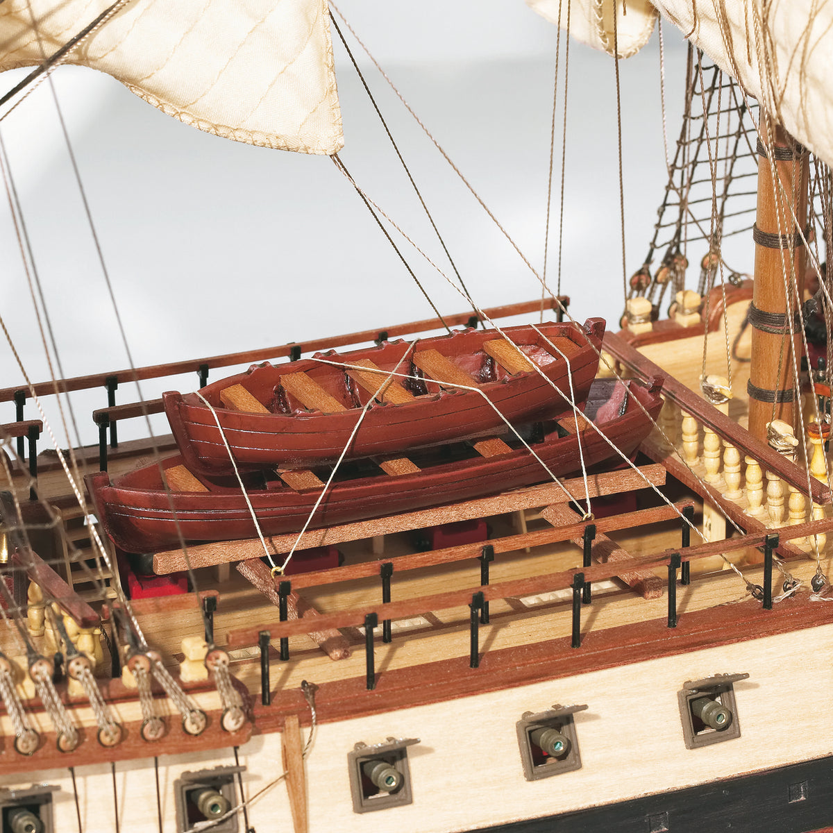 Close-up of a detailed wooden model ship deck with two small reddish-brown boats on the railing, cream-colored sails, rigging, and a light wood hull against a pale background.