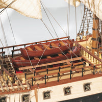 Close-up of a detailed wooden model ship deck with two small reddish-brown boats on the railing, cream-colored sails, rigging, and a light wood hull against a pale background.