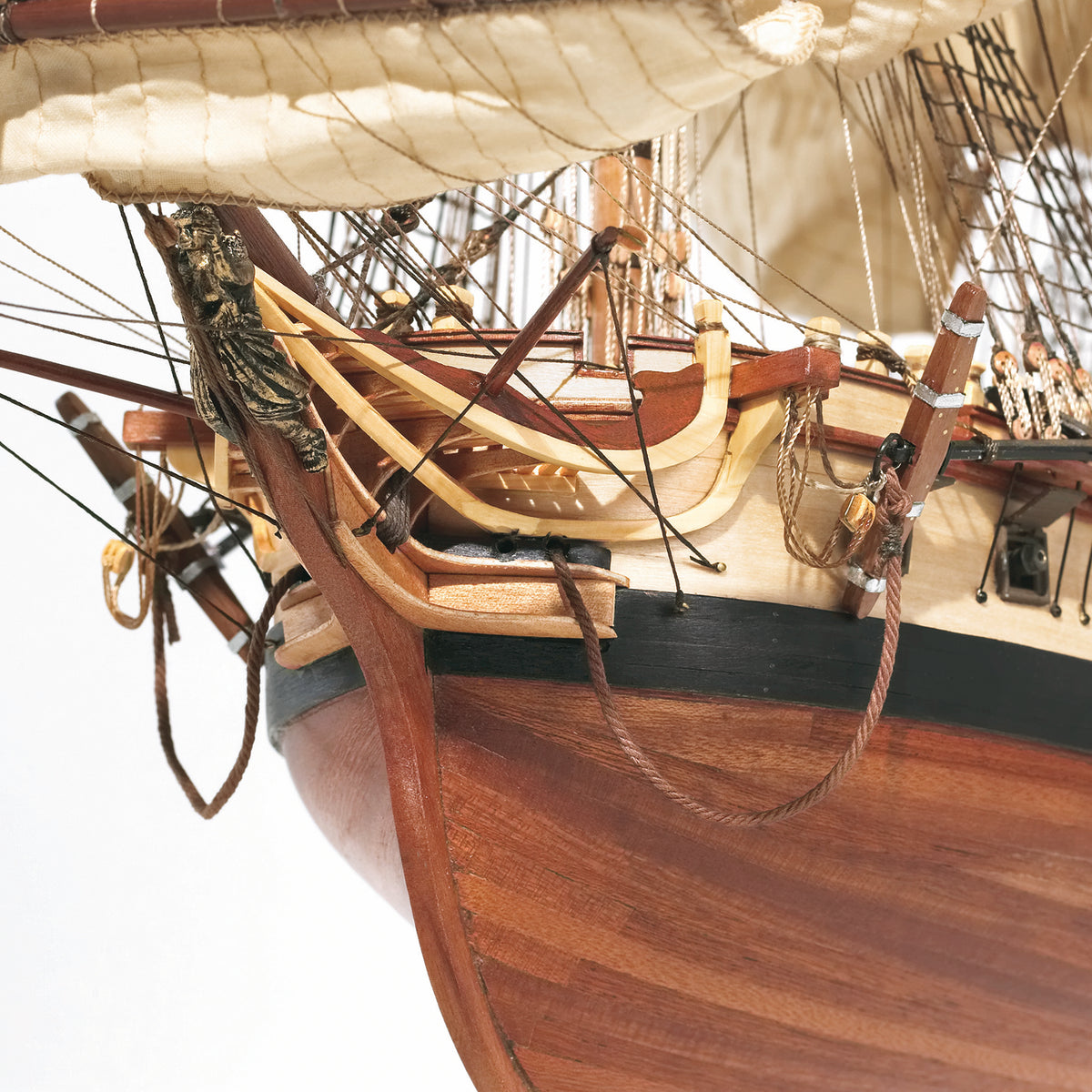 Close-up detail of a wooden model or replica ship, showing the hull, rigging, ropes, and part of the mast against a white background.