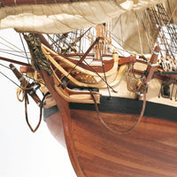 Close-up detail of a wooden model or replica ship, showing the hull, rigging, ropes, and part of the mast against a white background.