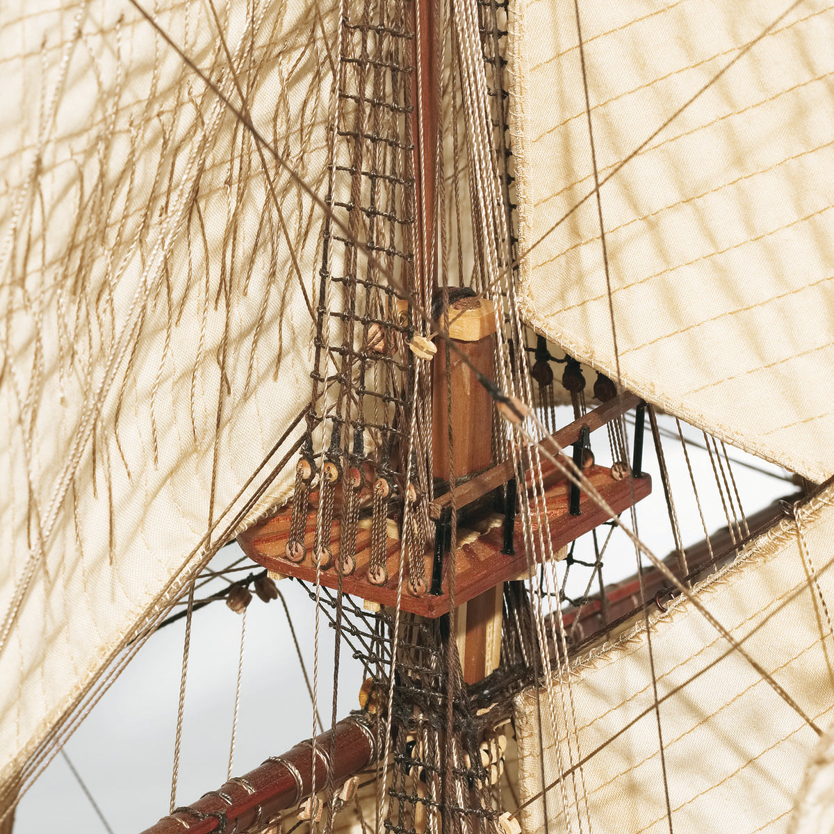 Close-up detail of a wooden sailing ship's mast, rigging, and large off-white canvas sails against a pale sky.