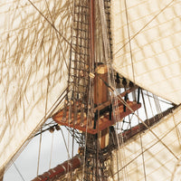 Close-up detail of a wooden sailing ship's mast, rigging, and large off-white canvas sails against a pale sky.