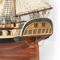 Close-up detail of a wooden model ship's bow and rigging, showing carved wood, white sails, brass chains, and decorative trim against a plain light background.