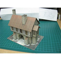 A miniature model house with a brown tiled roof, gray stone walls, and two chimneys, displayed on a green cutting mat.