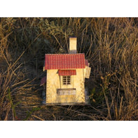 A miniature model of a cottage with a red tiled roof and beige walls, placed outdoors among dry grass.