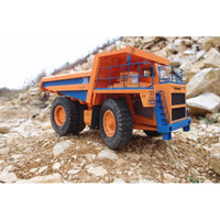 An orange and blue toy dump truck model with large black tires, positioned on a rocky, gravelly surface with a pile of rocks in the background.