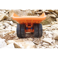 An orange toy dump truck with black wheels, photographed from the rear on a background of artificial or natural-looking rocks and gravel.